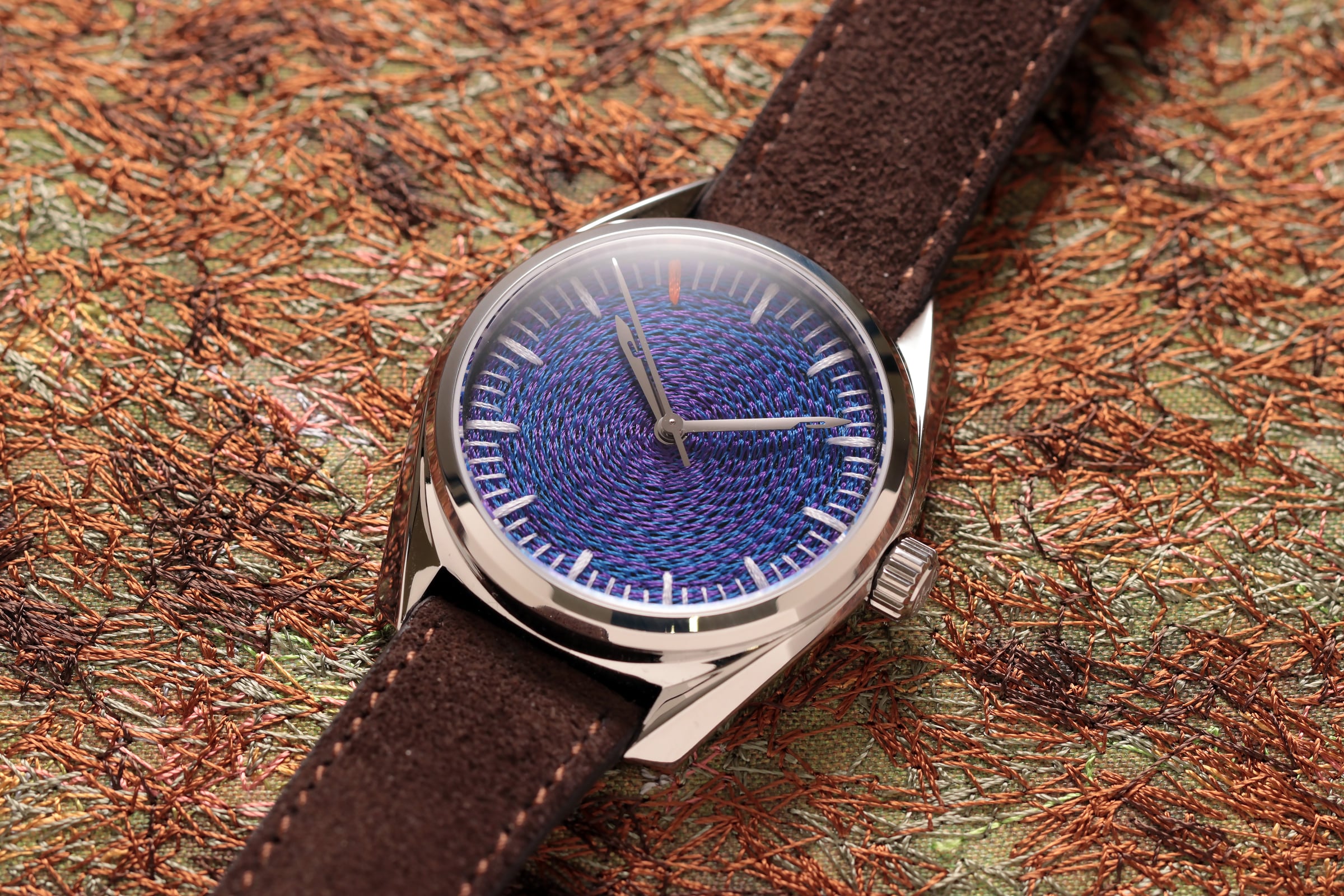 CORNELY wristwatch with a purple and blue spiral embroidered dial and a brown suede strap, resting on orange embroidered fabric