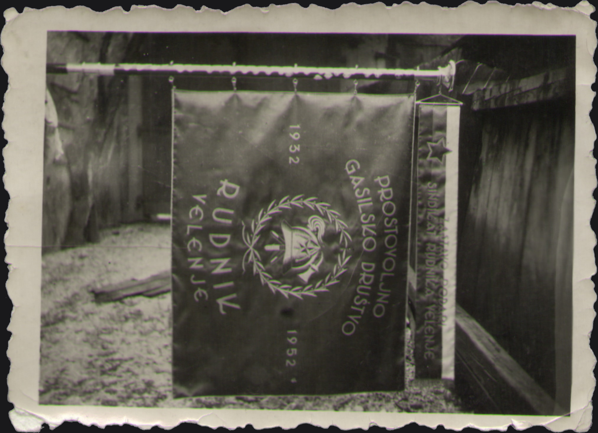Black-and-white photograph of an embroidered banner of the volunteer fire brigade Rudnik Velenje hanging from a pole, with a laurel wreath and the years 1932 and 1952