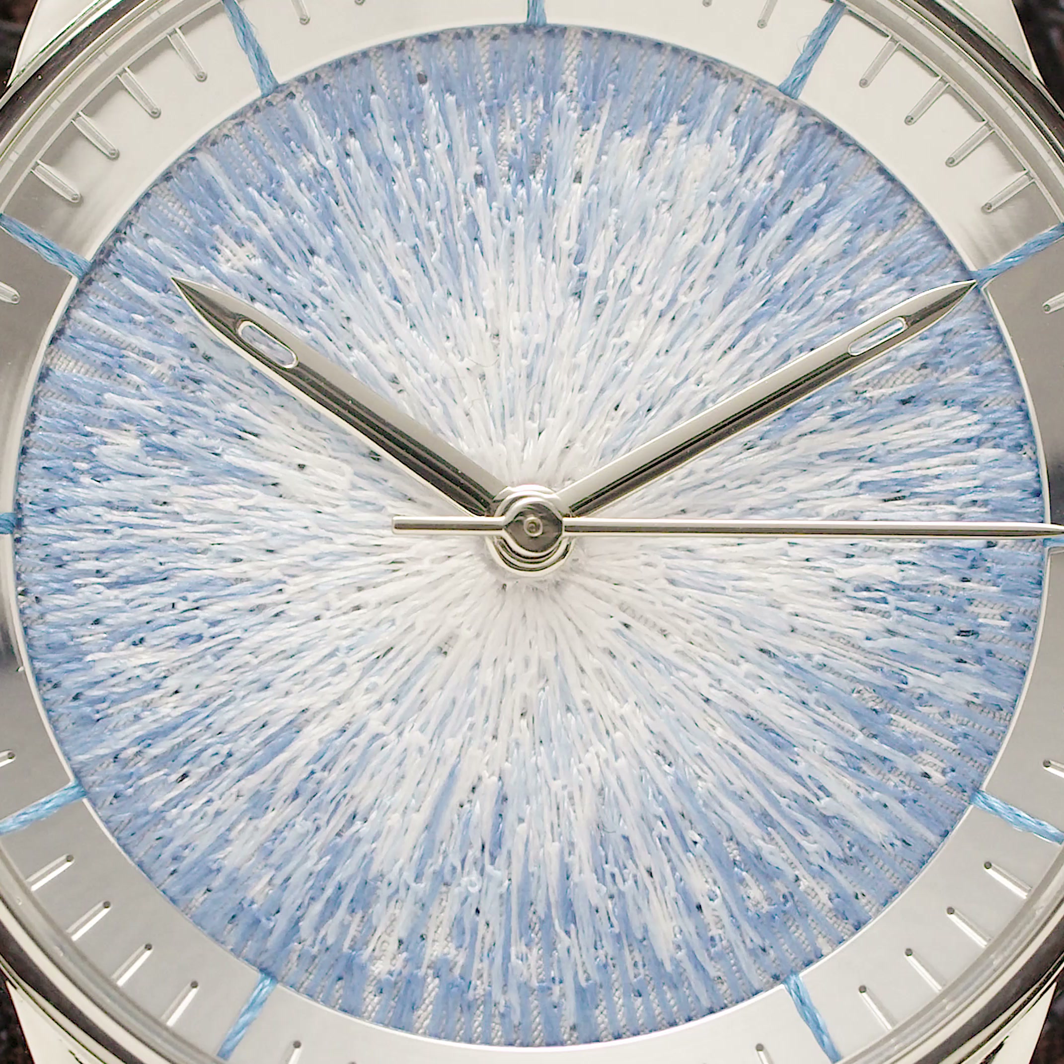 Close view of the JACQUARD dial: ice-blue sunburst stitch under a sloped chapter ring, needle-shaped steel hands across it